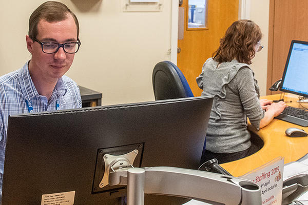 Two members of staff sit behind a curved wooden desk, each is looking at a desktop computer