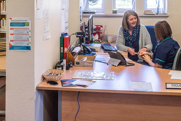 Two people sit behind a desk talking, one is dressed in an NHS nurse uniform. To the left the corner of the reading room can be seen with a table and bookshelves