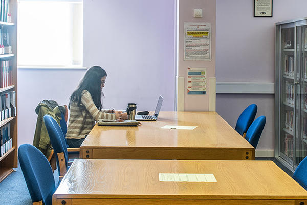 A student sits working at a large table, there are bookshelves either side